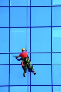 Ein Facharbeiter hängt an einem Seil in der Luft und reinigt eine große Fensterfront. (Gebäudereiniger)
