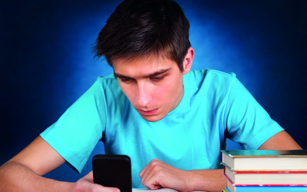 Ein Junge sitzt an einem Tisch, auf dem Bücher gestapelt sind und bedient sein Smartphone (Digitale Medien).