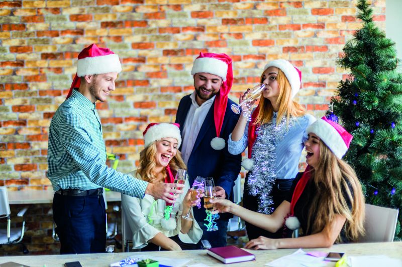 Fünf Arbeitnehmer tragen eine Weihnachtsmütze und stoßen lachend mit Sektgläsern an (Weihnachtsfeier im Unternehmen).
