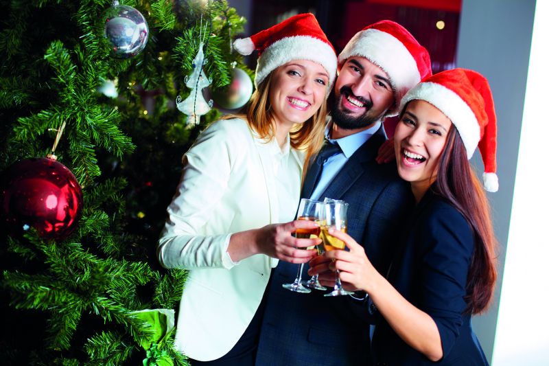 Drei gut gekleidete Arbeitnehmer stehen gemeinsam vor einem geschmückten Tannenbaum, lachen und stoßen mit Sektgläsern an (Weihnachtsfeier im Unternehmen).
