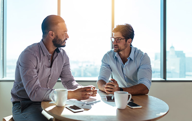 Zwei Männer sitzen gemeinsam an einem Tisch, trinken Kaffee und sprechen miteinander (Konflikte am Arbeitsplatz)