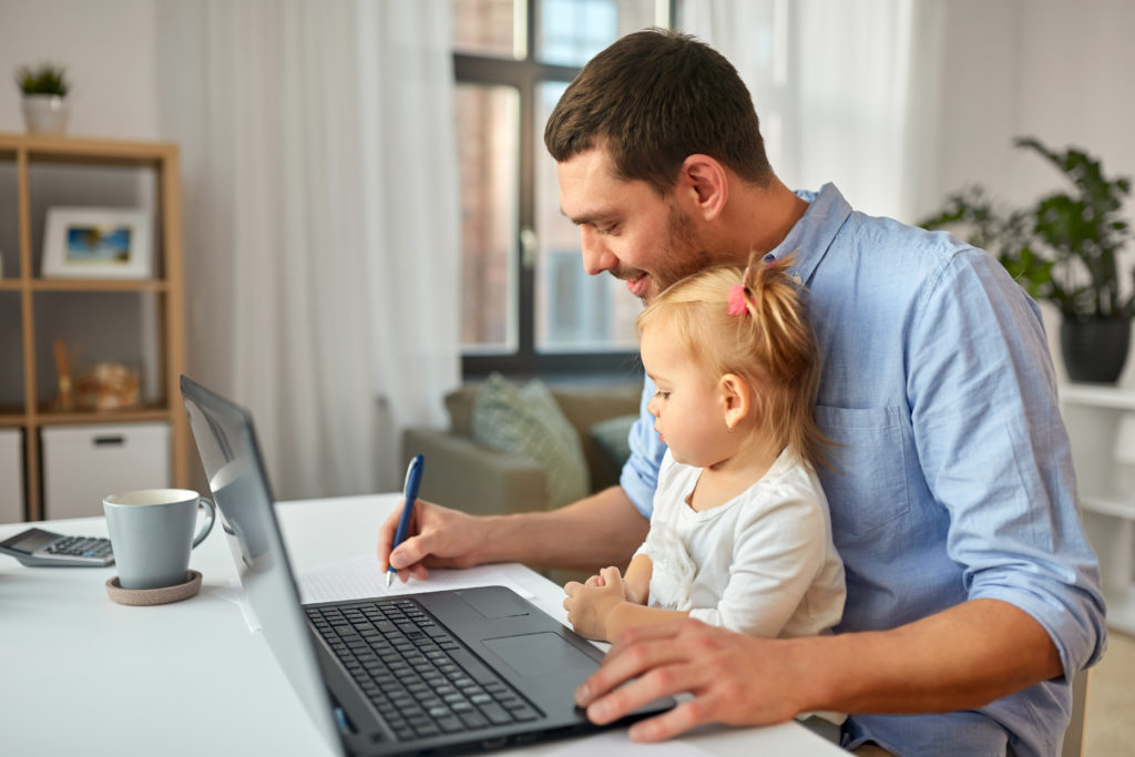 Arbeitnehmer und Corona: Ein Mann sitzt am Tisch und arbeitet am Laptop, während ein kleines Mädchen auf seinem Schoß sitzt.