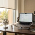 Hitze am Arbeitsplatz: ein kleiner Ventilator auf dem Schreibtisch und ein Glas Wasser an einem sonnigen Bürotag